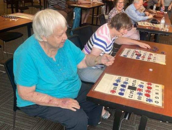From left to right, a woman wearing a blue blouse sits at a table next to a woman wearing a striped t-shirt. Each woman has a large-print Bingo card on the table in front of her.