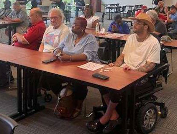 During a Bingo game, from left to right, a blind man wearing a red shirt sits at a table next to a man with a beard and glasses who sits next to a woman wearing a blue blouse who in turn sits next to a man wearing a straw hat who sits in a power wheelchair. The four people face toward the front of a room (not in the image). A large-print Bingo card is on the table between the woman in the blue blouse and the man wearing a straw hat.