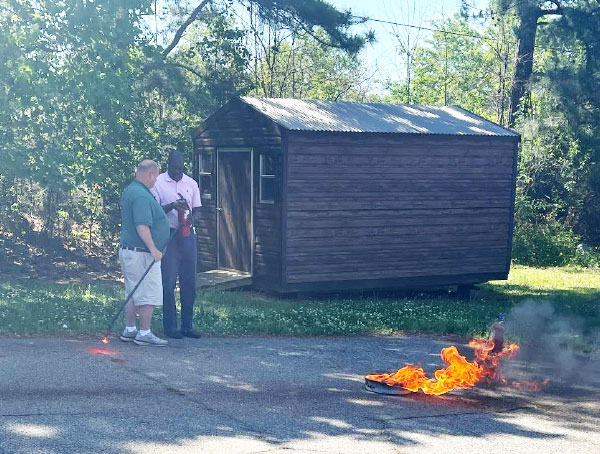 Two men stand in a parking lot at a safe distance from a small, contained, intentionally lit fire. The man in the white shorts instructs the blind man in the pink shirt in how to extinguish the fire.